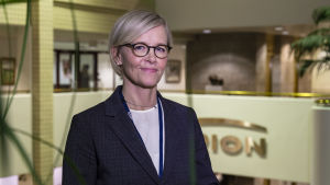Liisa Hurme ansvarar för leveranskedjan inom Orion. Liisa Hurme, en medelålders kvinna i blå kostym står på en balkong innuti ett hus. På väggen mittemot syns texten Orion.