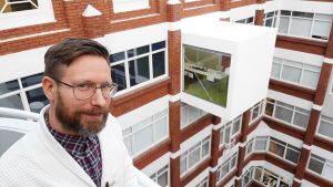 Nikolas Salomaa. Nikolas Salomaa, en man med glasögon, mustasch och skägg, står på en balkong mellan höga hus.