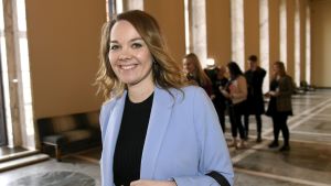 Katri Kulmuni är Centerns partiordförande. Katri Kulmuni har blont hår och en ljusblå kavaj och tittar leende in i kameran samtidigt som det ser ut som att hon är påväg någonstans.