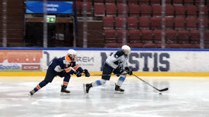 Ilona Palin åker ifrån en motståndare i en match mot Esbolaget Kiekko-Vantaa Itä. En ishockeyspelare åker skridsko med pucken vid klubbladet.