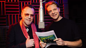 Pertti Neumann ja Olli Riipinen seisovat molemmin puolin katosta roikkuvaa mikkiä äänitysstudiossa hymyillen.