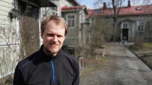 Oskar Lindroos, en man med ljust hår och ljust skägg, står på en innergård med gröna trähus.