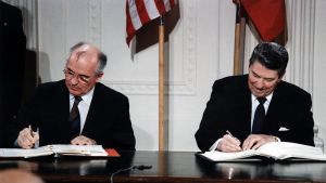 Gorbatshov ja Reagan istuvat pöydän takana ja allekirjoittavat sopimusasiakirjan kappaleita. Miehillä on mustat puvut ja kravatit. Taustalla seinällä ovat maiden liput.