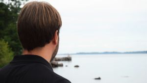 Från den södra stranden kan man skymta Helsingfors i fjärran. Tomi Ståhlberg står och ser ut mot havet. Han står med ryggen mot kameran. På sig har han en svart pikétröja.