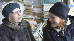 Glada grannar. Regissören Vilja Autiokyrö (t.h) hjälper gärna grannen Sinikka. Sinikka och Vilja sitter och skrattar mellan tidningshögarna.