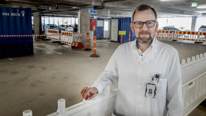 Antti Hakanen ser stora fördelar med att ha coronatestningen under tak i ett parkeringshus. Antti Hakanen, en man med mustasch och grånande skägg, står iklädd en läkarrock i ett parkeringshus.