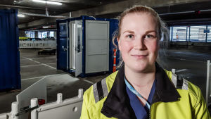 Mikaela Lahtinen trivs med sitt jobb. Mikaela Lahtinen, en ung dam med det ljusa håret i hästsvans, står iklädd en gul reflexväst framför en blå container där man tar coronaprover.