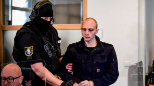 Den misstänkte gärningsmannen till attacken i Halle. Till höger en man i en svart jacka. Mannen har kort hår och är ganska liten. Till höger en polis som håller i honom i armen. Polisen är klädd i svart med hörlurar och mössa.
