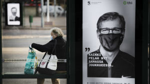 En skylt som föreställer en man i munskydd, bredvid sitter en kvinna på en bänk, också hon med munskydd.