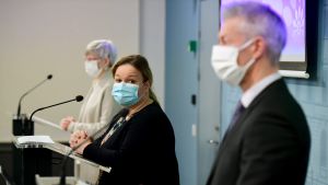 Kanslichef Kirsi Varhiala, familje- och omsorgsminister Krista Kiuru samt överläkare Taneli Puumalainen under torsdagens presskonferens. Tre personer står vid mikrofoner klädda i munskydd. I förgrunden en man, bakom honom två kvinnor.