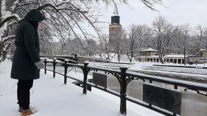 Två minnesplattor, en på svenska och en på finska, berättar att här vid Lilltorget i Åbo verkade Aurorasällskapet, som grundade Finlands första tidning. En dam i svarta vinterkläder tittar på två järnskyltar för Aurorasällskapet som hänger på ett staket vid Aura å. I bakgrunden syns Åbo Domkyrka och restaurang Pinella i ett snöigt Åbo.