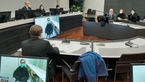 Den åtalade kvinnan deltog i rättegången via en videolänk från fängelset, eftersom hon exponerats för covid-19, och inte fick delta i rättegången på plats. Rättegångssalen vid en mordrättegång. Rättens ordförande och två tingsdomare sitter bakom ett långt bord uppe på en avsats, åklagaren och försvarsadvokaten sitter vid varsitt bord. Den åtalade är med på rättegången via en videolänk från fängelset.
