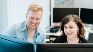 Mies ja nainen katsovat tietokoneen kahta ruutua hymyillen. Kuva on otettu tietokoneen näyttöjen takaa.