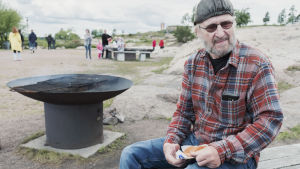Isto Arponen on eläkkeellä ja kertoo ulkoiluttavansa koiraansa usein juuri täällä. Tänään karvainen kaveri sai jäädä kotiin jotta Isto saisi syödä grillimakkaransa kaikessa rauhassa. Lakkiin ja ruutupaitaan pukeutunut parrakas mies istuu penkillä pidellen makkarapakettia käsissään.