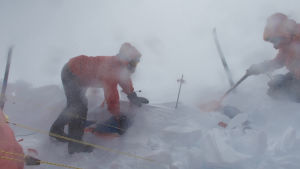 Att gräva fram en pulka mitt under pågående storm är inte lätt. Snön blir hård som cement. Två personer iklädda röda tjocka jackor och byxor. De befinner sig i ett snölandskap och det är storm. De gräver fram en pulka.
