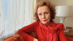 Klikkaa kuvaa ja lue lisää! Säveltäjä Kaija Saariaho istuu sohvassa ikkunan ääressä pukeutuneena oranssinpunaiseen huiviin ja puseroon