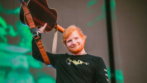 Ed Sheeran esiintymässä Helsingissä Malmin lentokentällä heinäkuussa 2019. Laula pitää akustista kitaraa päänsä takana ja hymyilee kohti yleisöä.