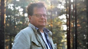 Arkeolog Jan Fast leder utrgrävningarna för projektet "Hangö 1941" som pågår till 2025. Man står i skogen. Solen skiner.