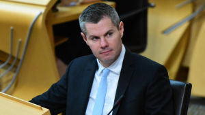 Derek Mackay är också medlem av det skotska parlamentet, där många ledamöter nu kräver att han lämnar sin plats. Derek Mackay, en medelålders man i blå kostym och kort grått hår sitter i sal med många stolar.