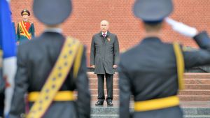 Sotilaat tekevät kunniaa Putinile, joka seisoo harmaassa päällystakissa portailla. Takana näkyy Kremlin punainen muuri.