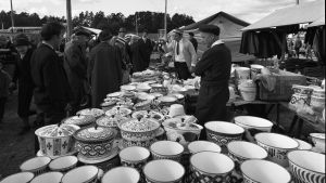 Emaljskålar till salu på Ekenäs höstmarknad. Svartvit bild av en försäljare på en marknad. På bordet står olika slags emaljskålar.