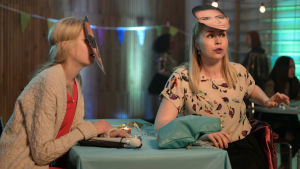 Donna (Alina Tomnikov) ja Mira (Essi Hellén) juhlissa naamiot kasvoillaan. Pöydällä Donnan keppi ja taustalla viirejä ja muuta juhlaväkeä.