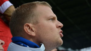 Johan Liljeström ropar: "Skjut!" En man med blå halsduk och ljusblå jacka hejar på sitt lag på en fotbollsmatch. Läpparna formas till ordet "Skjut!".