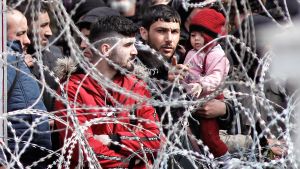 Syriska flyktingar på gränsen mellan Turkiet och Grekland. Joukko Kreikkaan pyrkiviä turvapaikanhakijoita seisoo piikkilanka-aidan takana. Kuvan keskellä on parrakas mies, joka pitelee sylissään pikkulasta ja osoittaa sormellaan kuvaajan suuntaan.*
