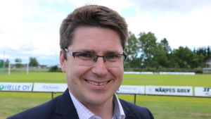 Mikko Ollikainen. En man med mörkt kort hår och glasögon. I bakgrunden syns en fotbollsplan.