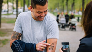 Tatueringar blir allt vanligare. Här har någon tatuerat Anders Tegnell på armen. Mies näyttää käsivarrestaan tatuointia kännykkäkameralla kuvaavalle naiselle.