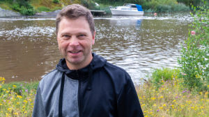 Sampo Vainio hoppas att öringen också leker i Tjusterbyforsen om tio år. En leende man vid en å. Vit motorbåt ankrad i bakgrunden.