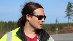 Pekka Pyyny från Neoen ser stor potential för vindkraften i hela Kustösterbotten. En man med solglasögon och långt mörkt hår i en hästsvans står i en skogsöppning.