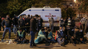 Människor väntar på gatan i Bayraklidistriktet i Izmir. Människor sitter på en trottoar. De bär munskydd och en del är insvepta i filtar. I bakgrunden en ambulans.