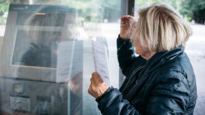 Eläkeikäinen nainen pankin ulkupuolella lasku kädessä.