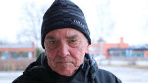 Lars Österblad. En i 70-årsåldern med en svart mössa på huvudet står på en parkeringsplats.