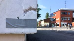 Fokushusets kondition granskades i vintras. En marmorvägg där en bit marmor lossnat och den har tejpats fast med isoleringstejp. I bakgrunden syns gata, hus, cyklist. Blå himmel, sommar.