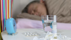 När man är förkyld eller har influensa ska man helst vila. I förgrunden finns mediciner och näsdukar på ett bord, i bakgrunden ligger en förkyld man i sin säng