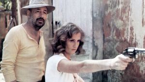 Philippe Noiret ja Isabelle Huppert. Auringonpimennys-elokuvassa Philippe Noiretin ja Isabelle Huppertin esittämät hahmot seisovat ulkona, nainen pitää asetta kädessään ja osoittaa sillä jotain.