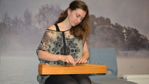 Ung kvinna med långt mörkt hår spelar på ett kanteleliknande instrument. Sitter på blå stol med bakgrundstapet som föreställer vintrigt landskap