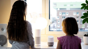 Smarta assistenter i hemmet. Två flickor i ca 6 och 9 årsåldern står vid ett fönster med ryggen till kameran och har två smarthögtalare framför sig som de talar till