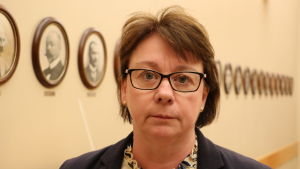 Åsa Blomstedt. En kvinna med kort mörkt hår och glasögon står intill en vägg som är prydd med porträttbilder.