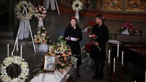 Ari Behns dotter Maud Angelica Behn håller tal på begravningen. Bredvid står prinsessan Märtha Louise av Norge – Behns ex-fru. Två kvinnor står vid vit kista som omges av flera blomkransar.