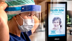 En sjuksköterska tar på sig skyddsutrustning - i bakgrunden ett köpcenter med en uppmaning att stanna hemma på en stor reklamskylt, text på svenska