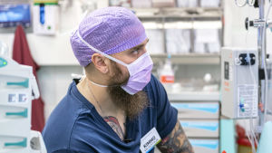 Parrakas mies työskentelee sairaalan leikkausalissa kasvomaski naamallaan.