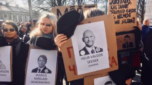Demonstranterna utanför Dombergets slott i torsdags hade plakat med politiker från Centerpartiet och Isamaa-Fäderneslandet. Hattarna symboliserar att de nu befaras styras av Ekres ledare Mart och Martin Helme. De bär alltid hatt. Demonstranter utanför Riigikogu i Tallinn håller upp skyltar med porträtt på politiker och en hatt ovanför som symboliserar att politikerna låter sig styras av Ekre-politikerna Mart och Martin Helme, som alltid använder hatt.