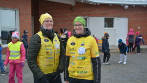 Heidi Martens Två kvinnor i gula västar utanpå sina vanliga kläder, barn i bakgrunden.