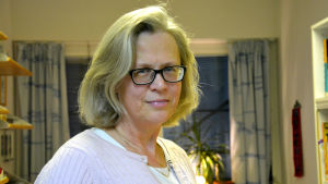 Revisionsnämndsmedlemmen Anne Eriksson tycker att man ska få kritisera tjänstemän, ordförande och nämndmedlemmar i revisionsnämnden även utanför nämnden och är kritisk till bilagan som säger annorlunda. en kvinna i ljust hår och ljusa kläder och mörka glasögon står mitt i ett kontorsrum