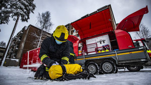 Det är ytterst viktigt att kontrollera sin tryckluftsflaska och sele inför rökdykningen En brandman med gul hjälm sitter på den snöiga marken och gör sin gula tryckluftsflaska färdig för rökdykning. Bakom honom står en röd brandkårsvagn med utrustning parkerad.