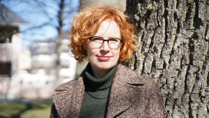 Sirkku Varjonen har en fot i det urbana men står ändå stadigt med båda fötter i rurala Bromarv. En kvinna med rött lockogt hår står ute lite lutad mot trädstam.