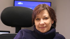 Maria Launonen. En kvinna i 50-årsåldern sitter i en kontorsstol. Bakom henne skymtar en tv-skärm.